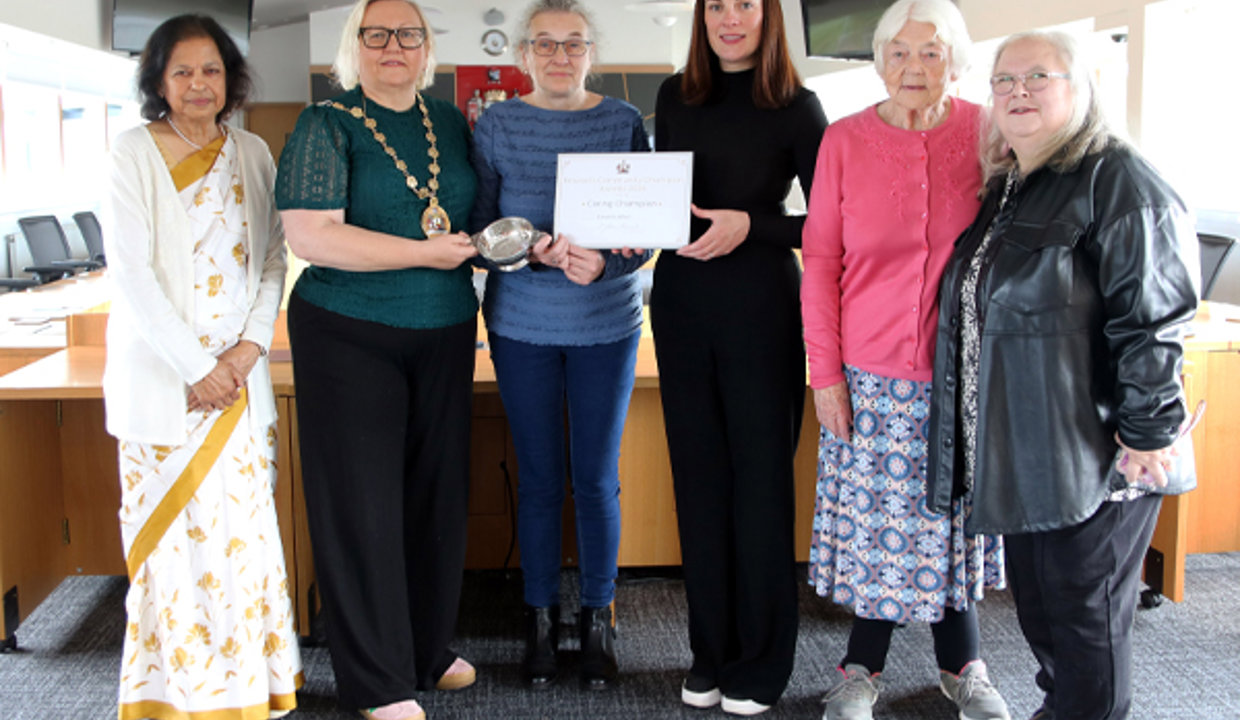 Formal shot of six women standing in Council chambers.  The women in the centre of the pic are holding a framed certificate and a quaich. 