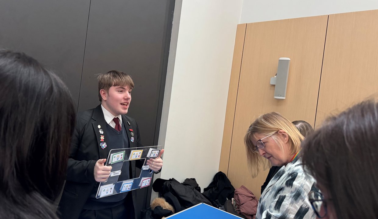 School boy wearing school uniform holding a Perspex learning tool in his hands standing behind a blue desk.  In front of him are women listening to him and looking at items on the desk. 