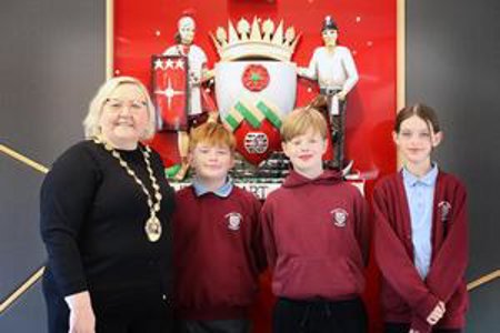 Provost Renwick, Ruairidh, Isaac and Sophia standing in front of East Dunbartonshire’s Coat of Arms which is mounted on the wall.  Crest has red background, with a shield in centre and soldier and a miner at either side of coat of arms. 