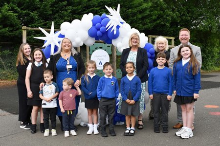 Children and adults standing outside in front of a large balloon arch, made up of blue and white balloons and large white stars. Most of the children are wearing royal blue school jumpers.