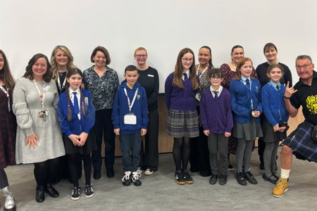 Group of adults and children wearing school uniforms standing in front of large white projector screen.  In the far right hand corner is a man wearing a kilt and sporran and a T-shirt which reads ‘Reading Rocks’ and he’s holding two books in his right hand. He’s lunging to the side and making a rock sign with his fingers on his left hand. 