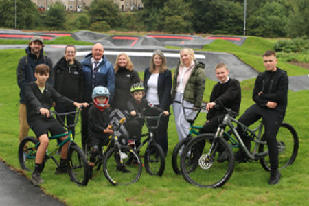 A group of people some standing and others on the bikes. Behind them is the pump track.