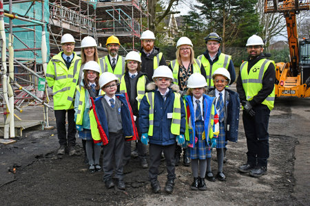 Group of children and adults wearing hi-viz jackets and white hard hats standing beside a building covered in scaffolding.