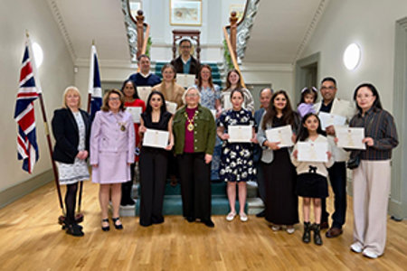 Twelve men, women and children holding certificates standing around a carpeted staircase.  The provost is standing in the middle of the picture. Both the Union flag and the Saltire are hanging from a stand on the left of the staircase.