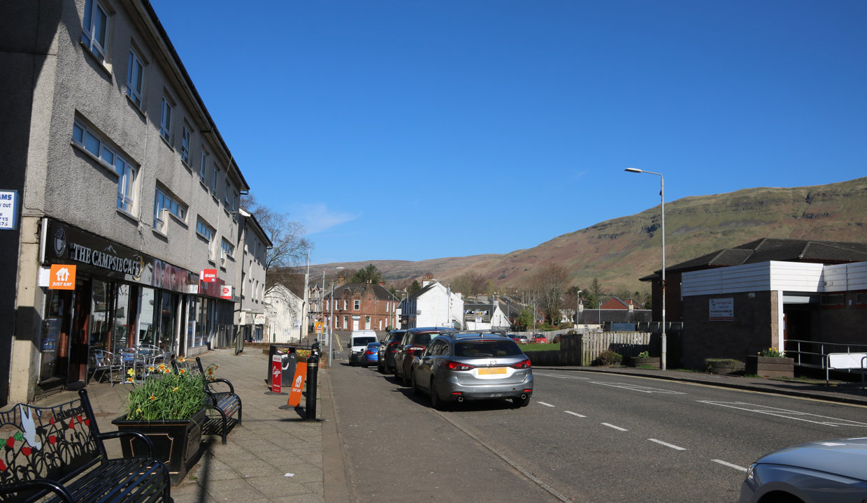 A view down the main street of Lennoxtown towards the Campsie Fells in the background. Cars are parked along the roadside and there is a row of shops with benches on the pavement in front of them and flats above. 