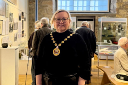 Woman wearing a black top and chains of office standing in large room with exhibits. A brick work wall and large window is behind her. 