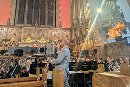 Inside of a church with large stained-glass window in background.  In the foreground is a band playing instrutments, led by a conductor with his back to the camera.  