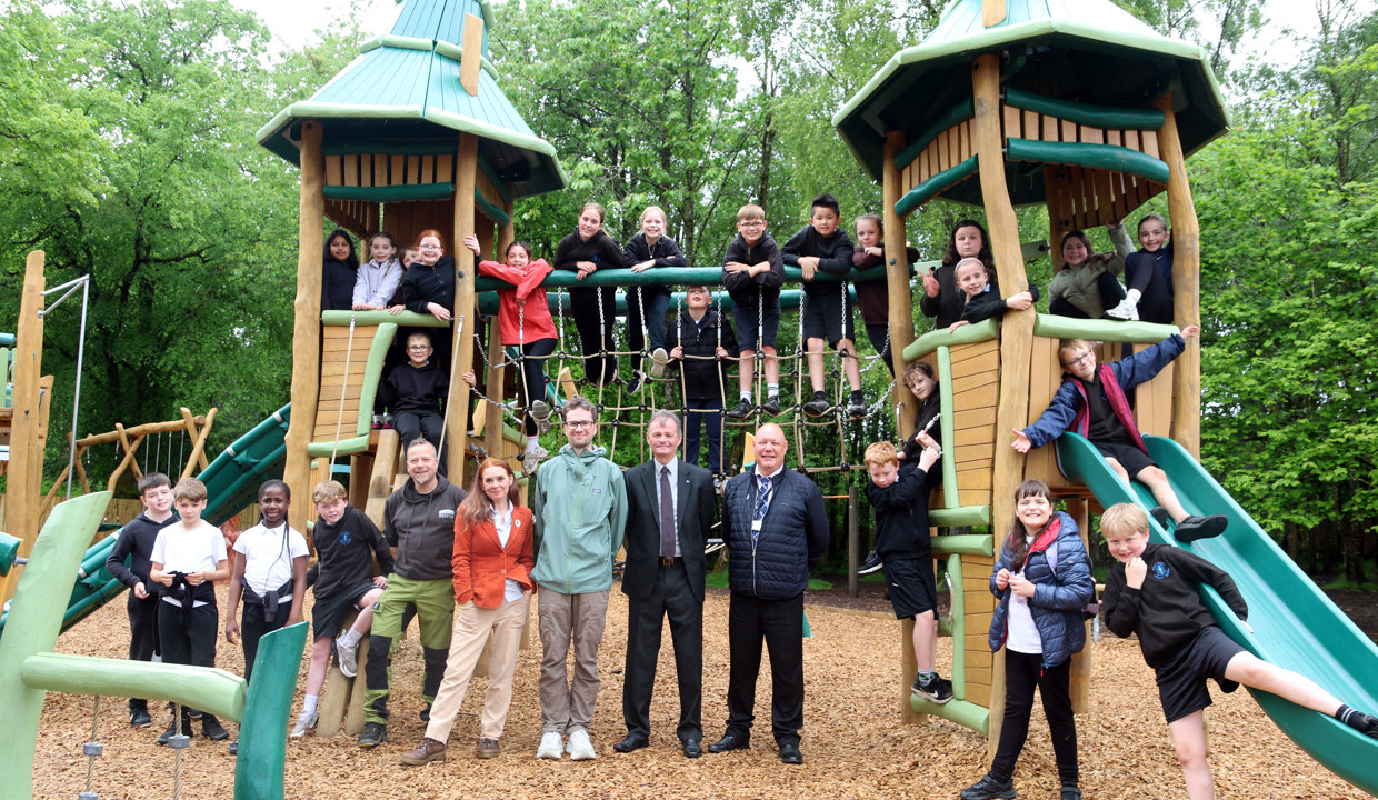 A group of around 20 school children pose on a climbing frame which has a rope bridge and two wooden turrets at either end. A group of five adults stands in the middle of the children. 