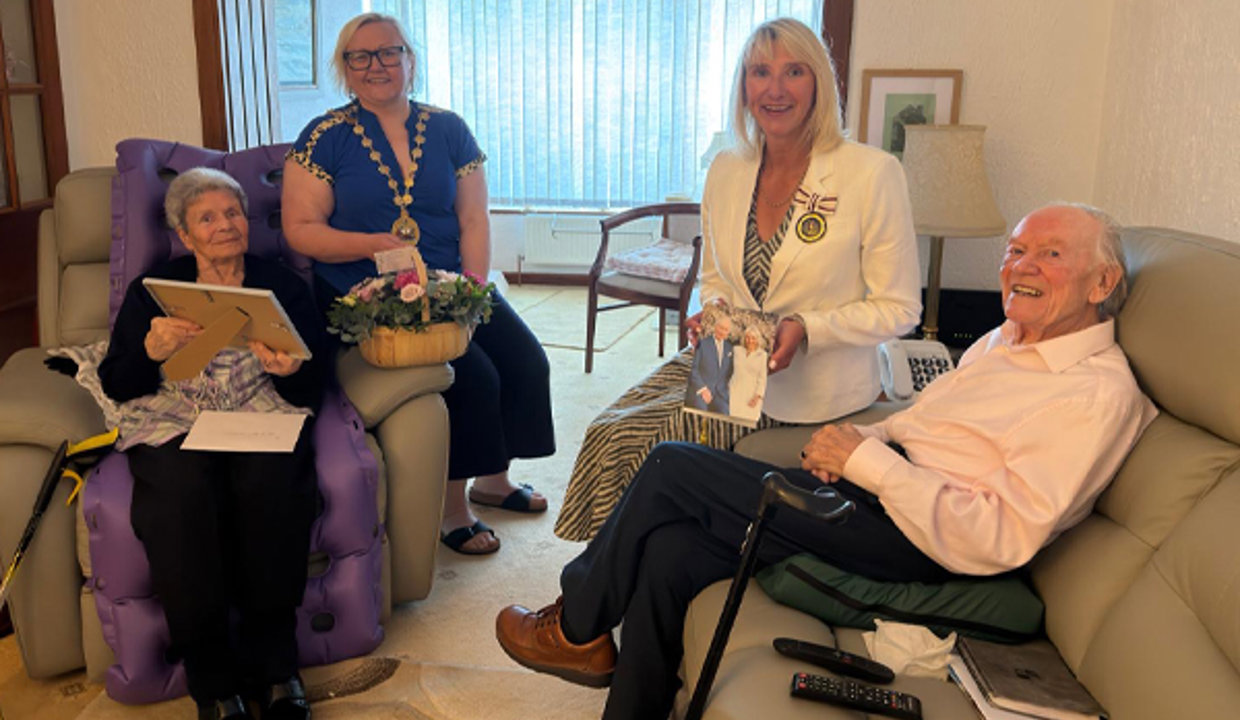 Picture of four people in a living room. On the left is a woman sitting in a chair holding a picture frame.  Next to her is a woman wearing a chain of office and holding a basket of flowers.  Across from them is a woman wearing a medal. She is holding an image of The King and Queen Camilla and she is sitting on the arm of a sofa beside a man.  All four are smiling at the camera. 