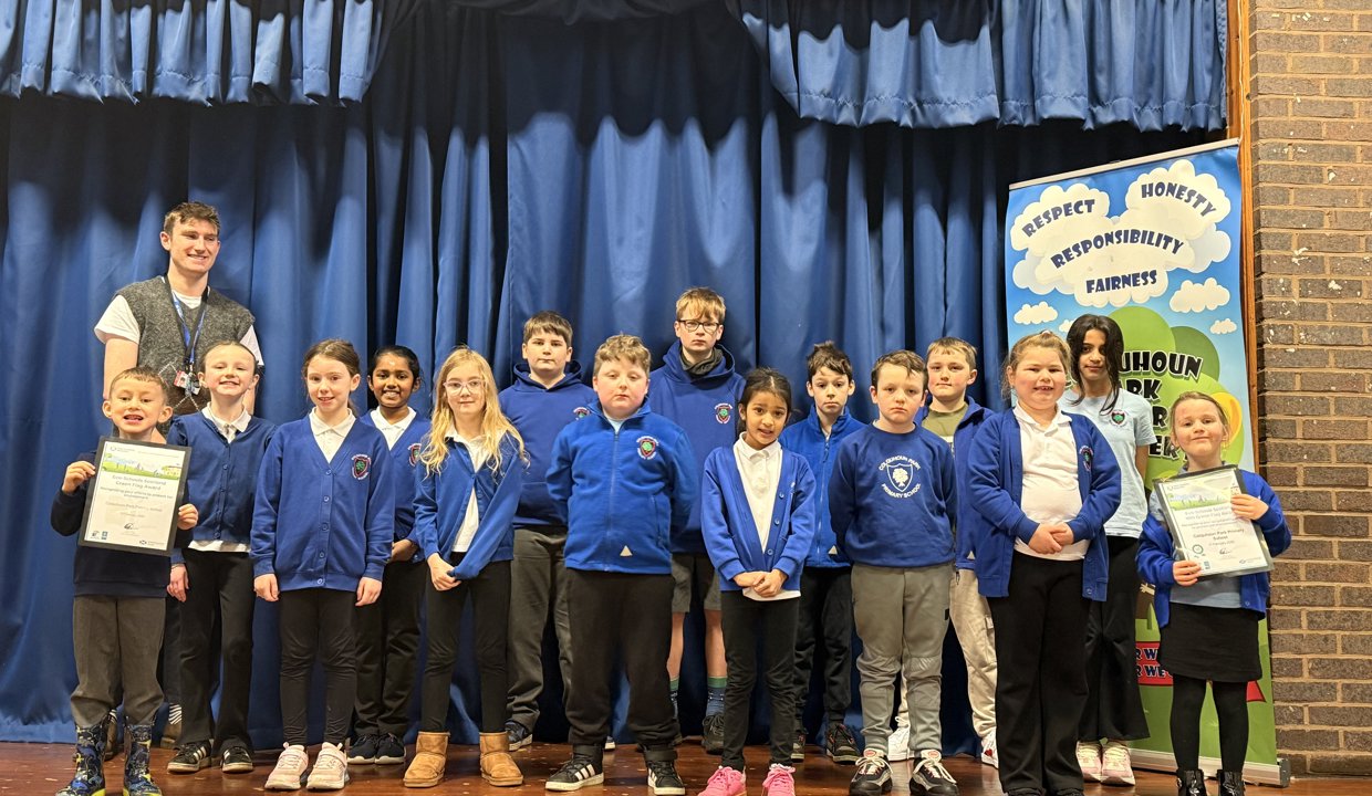 Group of children wearing blue school jumpers standing on a school stage. Behind them on the left is a male teacher wearing a grey pullover.  There is a blue stage curtain behind the group.