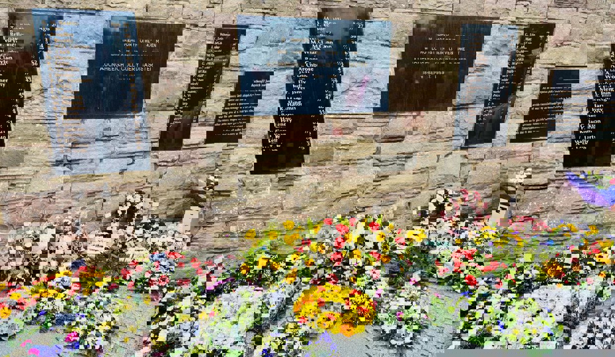 Beautiful, colourful flower arrangements laid against a sandstone-coloured wall with large black plaques interspersed along the brick work.  The plaques contain a list of names of those who died at the Auchengeich Mining Disaster in 1959.  