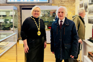 Woman wearing a black top and chains of office standing beside man wearing dark coloured coat and shirt and tie. Behind them is a brick work wall and large window
