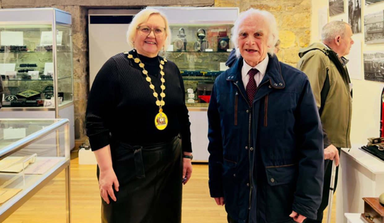 Woman wearing a black top and chains of office standing beside man wearing dark coloured coat and shirt and tie. Behind them is a brick work wall and large window