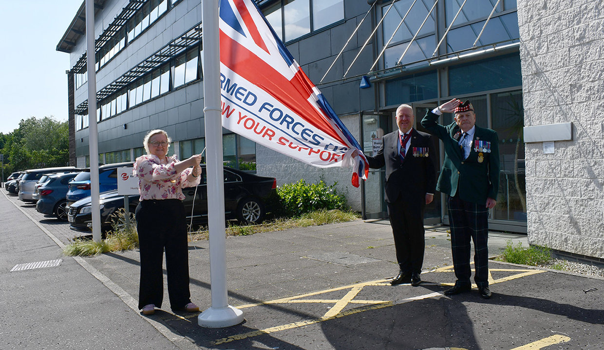 Provost Gillian Renwick, wearing chains of office raises special union flag – the Armed Forces Day Flag. Across from the flag pole stands a man in a suit wearing military medals and a man in military dress, wearing a hat and saluting the raising of the flag. 