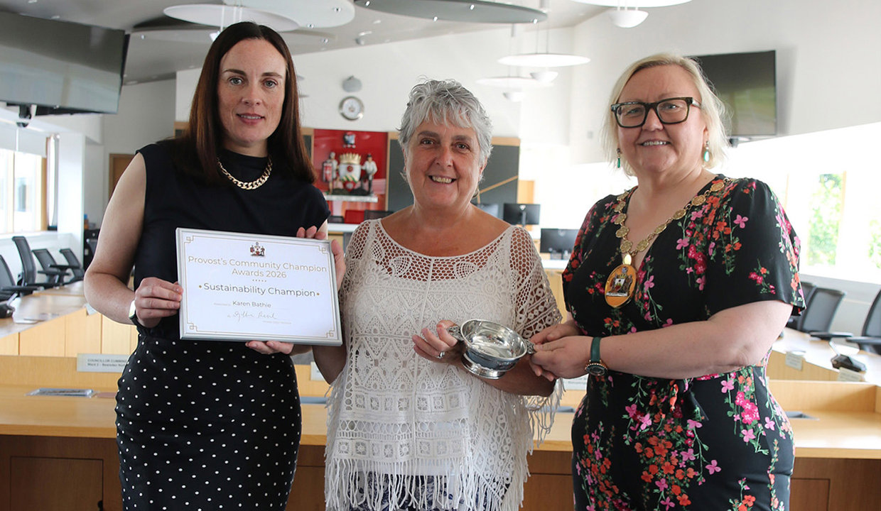 Three women standing together in council chambers.  Woman on left is holding framed certificate.  The woman in the centre is resting one hand on the fram and in the other she is holding a quaich.  The third woman to her right is also holding onto the quaich and she is wearing chains of office. 