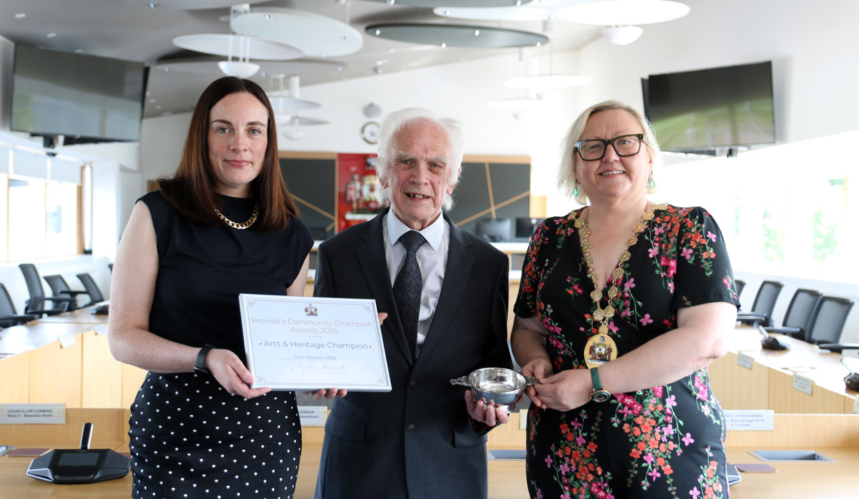 To the left is a woman wearing black, holding a framed certificate next to older gentleman wearing a suit. He is holding one side of a silver quaich and the other side is held by a woman wearing chains of office. 