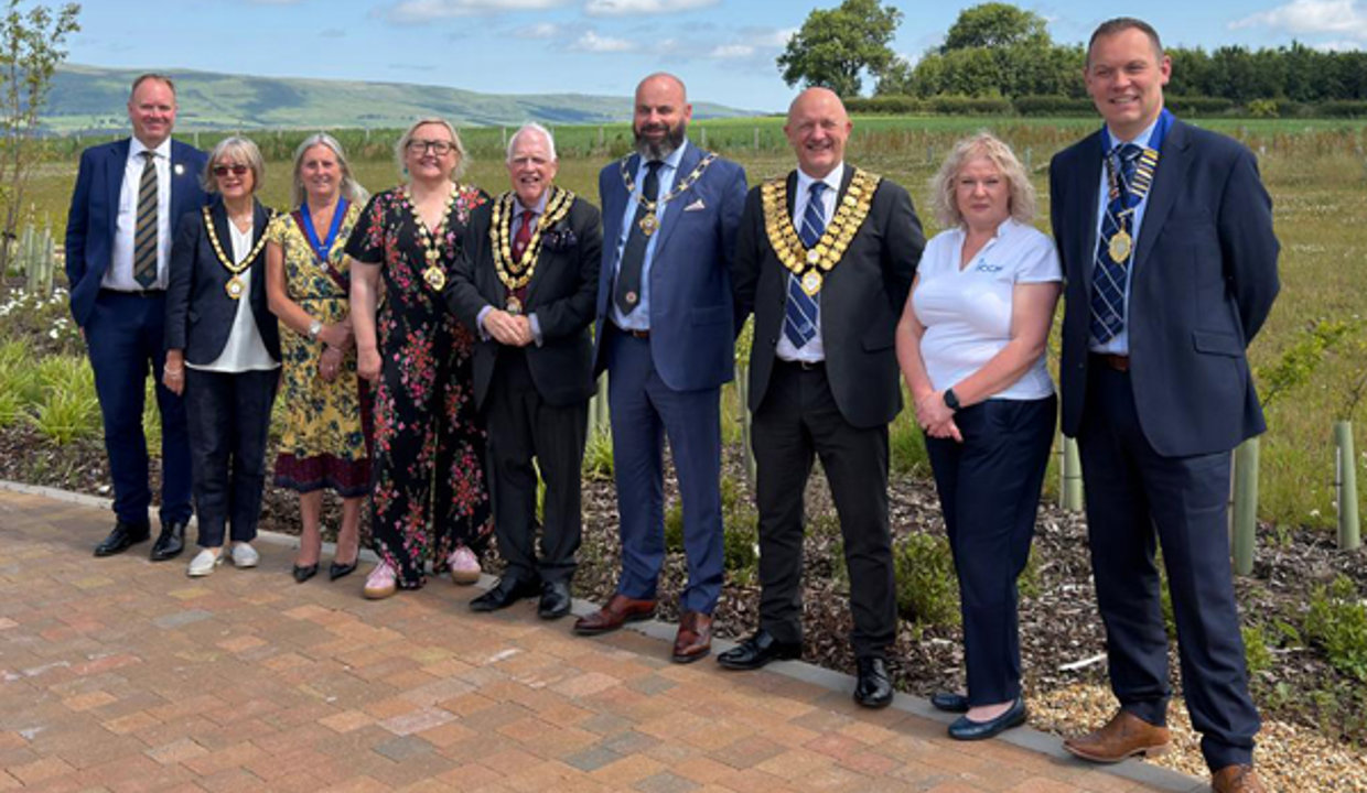 Nine people smartly dressed, some wearing chains of office, standing side by side on a monobloc path.  Behind them are green fields, the Campsie hills and a beautiful blue sky, with fluffy white clouds.