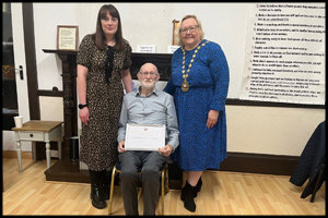 Man seated in centre holding certificate, surrounded by two women, standing either side of him.  The woman to his right is wearing the Provost’s Chains of East Dunbartonshire. 