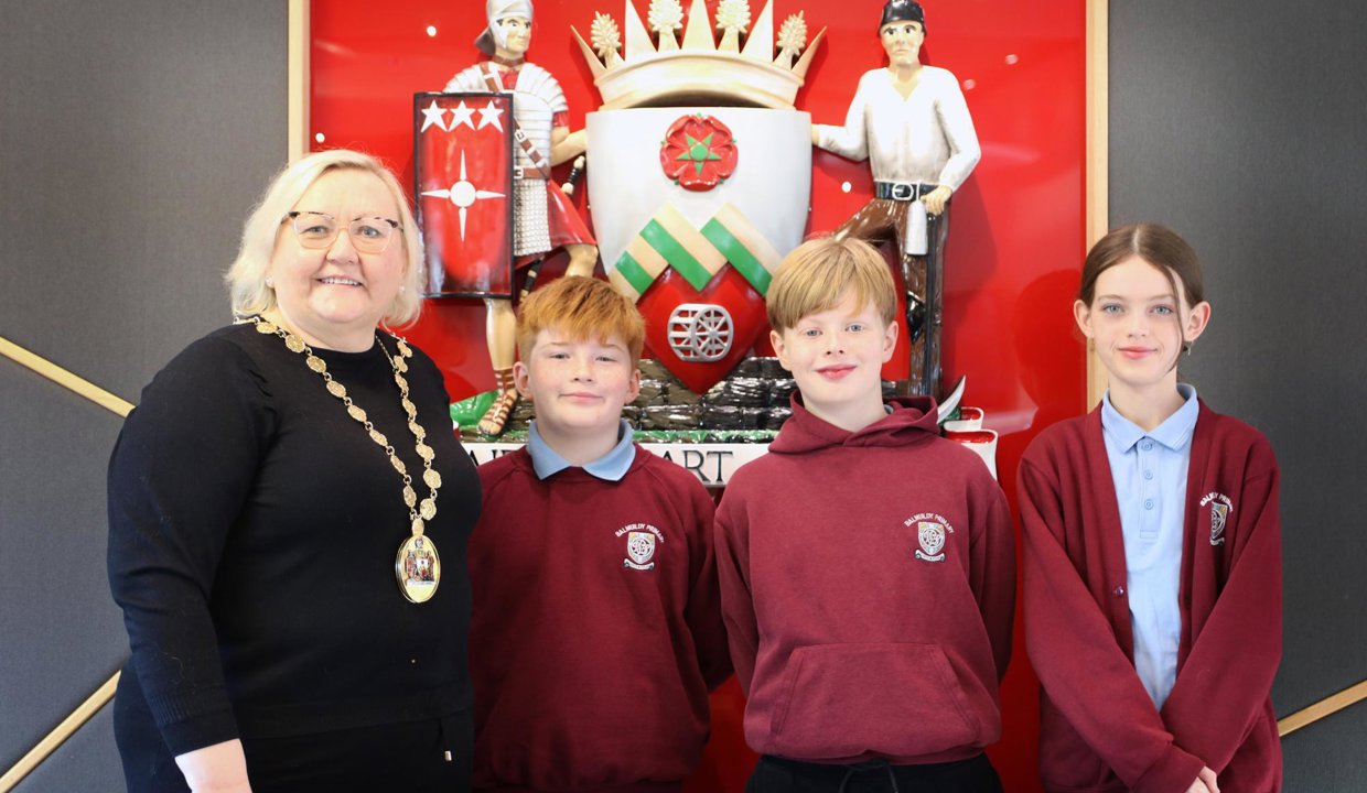 Provost Renwick, Ruairidh, Isaac and Sophia standing in front of East Dunbartonshire’s Coat of Arms which is mounted on the wall.  Crest has red background, with a shield in centre and soldier and a miner at either side of coat of arms.