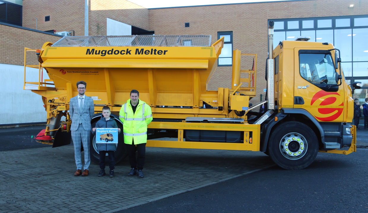Councillor Paul Ferretti and Fleet manager present winning pupil with a certificate in front of a yellow gritting truck. 