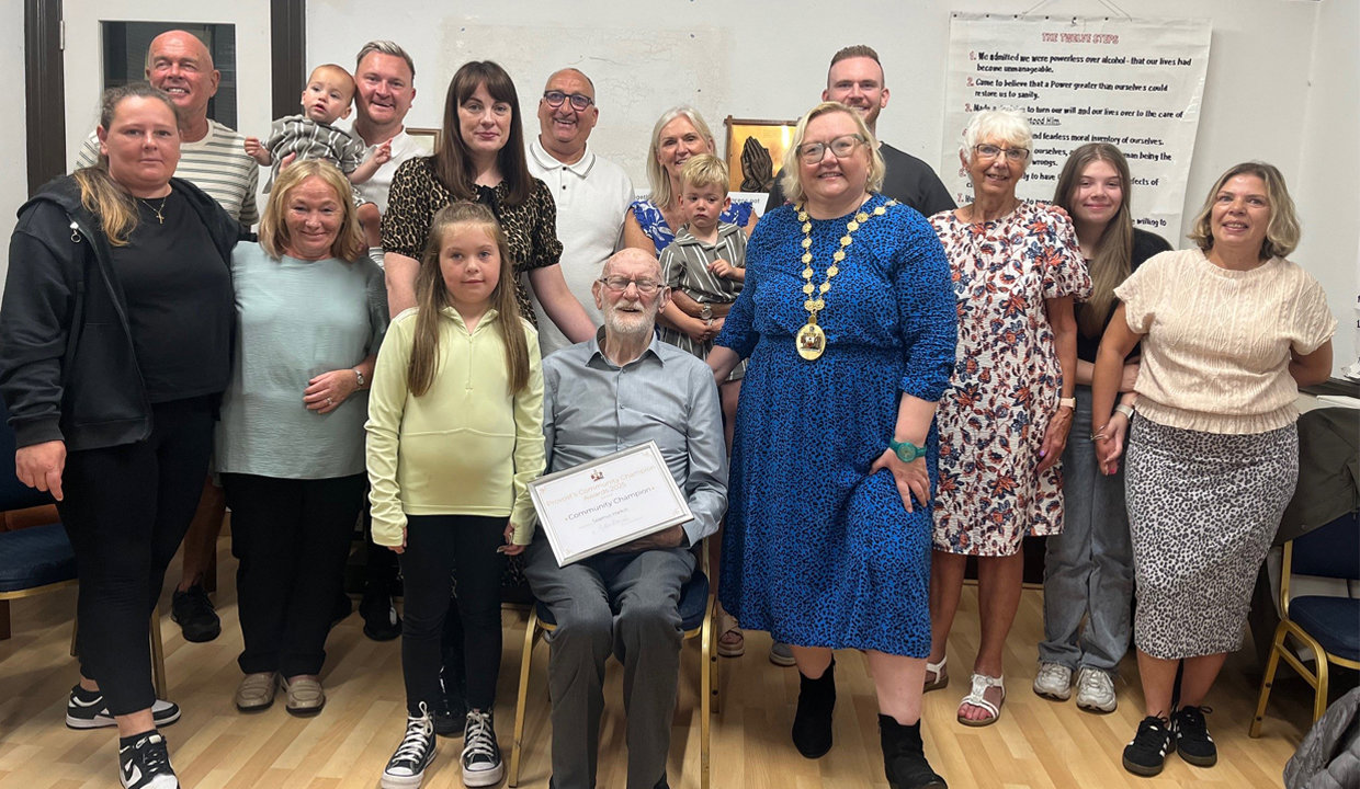 Man seated in centre of image holding a certificate, surrounded by men, women and children standing and smiling towards the camera. The woman to his right is wearing the Provost’s Chains of East Dunbartonshire.