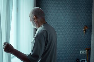 An elderly man looks out of a window after a knock at the door