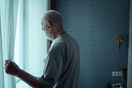 An elderly man looks out of a window after a knock at the door