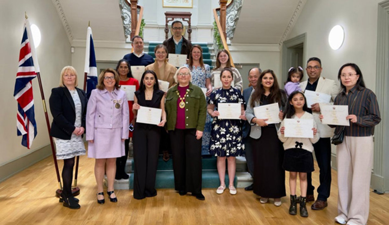 Twelve men, women and children holding certificates standing around a carpeted staircase.  The provost is standing in the middle of the picture. Both the Union flag and the Saltire are hanging from a stand on the left of the staircase.