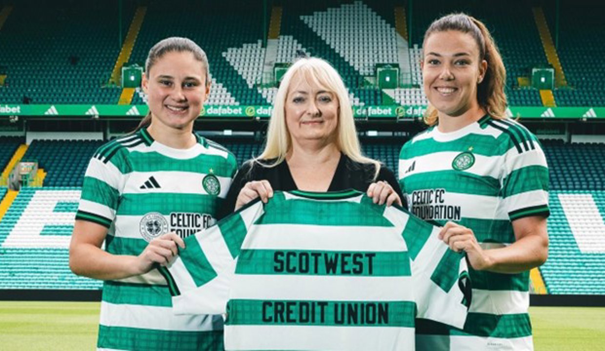Three women at Celtic Park. They are holding up a Celtic top that has Scotwest Credit Union printed on the back.