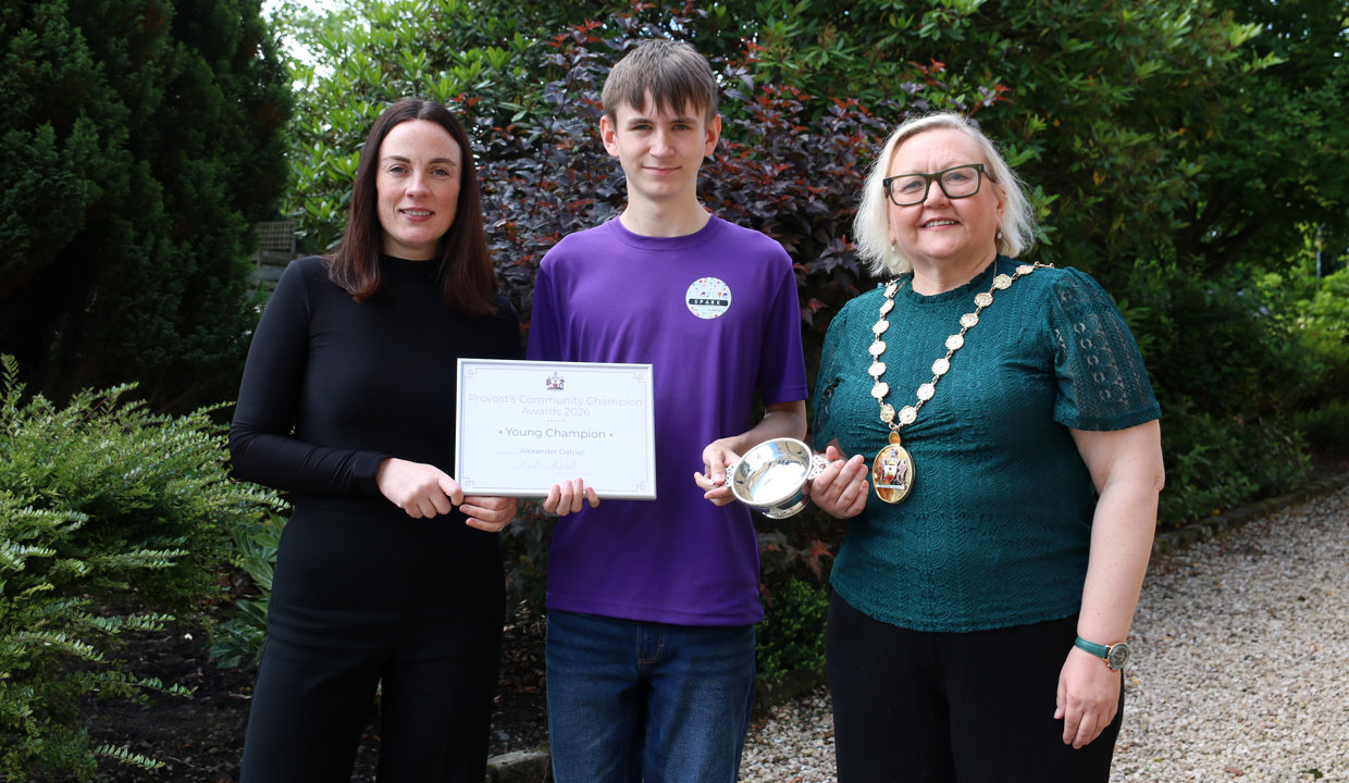 Young man in a purple top standing between two women – the one on the left wearing black and holding a framed certificate, the one on the right wearing a green top, a chain of office and holding a silver quaich. They are outside and there is green foliage behind them. 