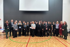 Large group of people standing in a gym hall with a large black wall behind them with a white screen on top. 