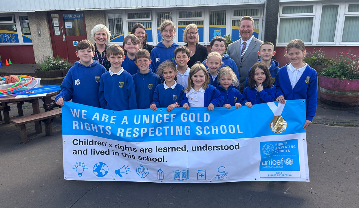 Four adults and a group of primary school aged pupils wearing royal blue school cardigans and jumpers standing  behind a UNICEF Gold Rights Respecting School banner. 