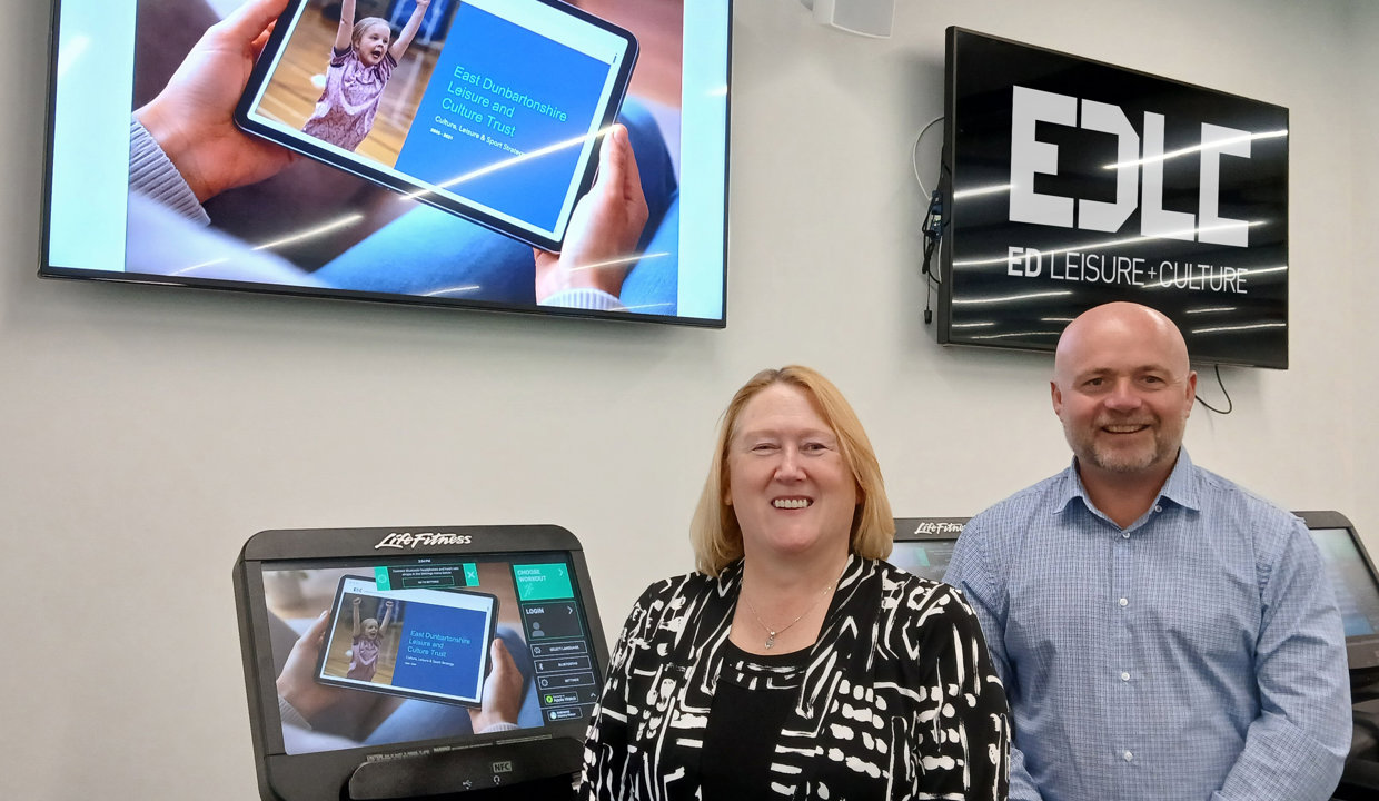Karin Jackson, General Manager of EDLCT and Alan Wright, Chair of EDLCT Board, are pictured at the launch of the Strategy StoryMap. They are standing beside an exercise machine displaying an image of the StoryMap, with TV screens behind.