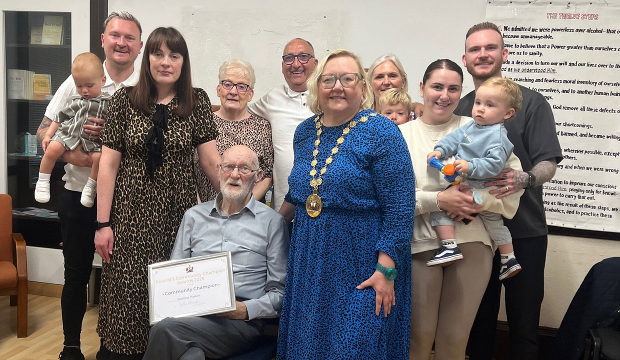 Man seated in centre of image holding a certificate, surrounded by men, women and children standing and smiling towards the camera. The woman to his right is wearing the Provost’s Chains of East Dunbartonshire.