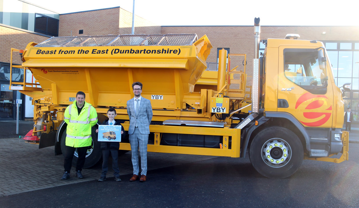 Councillor Paul Ferretti and Fleet manager present winning pupil with a certificate in front of a yellow gritting truck