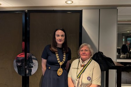 Two women facing camera – one wearing a blue dress and chains of office around her neck, the other wearing a beige scout uniform and necktie
