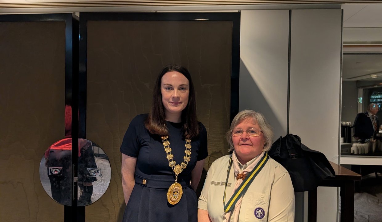 Two women facing camera – one wearing a blue dress and chains of office around her neck, the other wearing a beige scout uniform and necktie