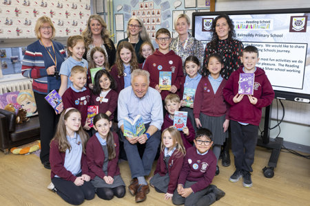 A group of children wearing burgundy school jumpers standing or sitting around a man smiling at the camera and reading a book. Five women are standing at the back of the group, smiling at the camera.