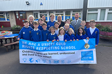 Four adults and a group of primary school aged pupils wearing royal blue school cardigans and jumpers standing  behind a UNICEF Gold Rights Respecting School banner. 