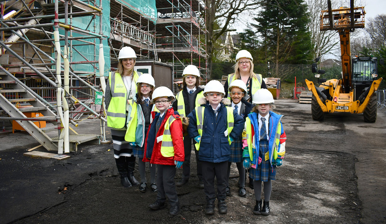 Group of children and adults wearing hi-viz jackets and white hard hats standing beside a building covered in scaffolding.
