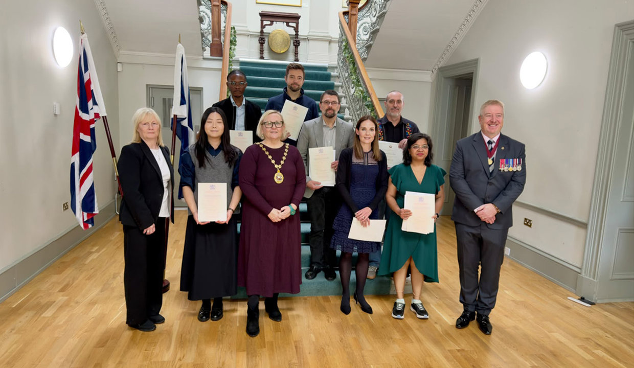 Group of people standing at the foot of a beautiful staircase with union and saltire flags on display at the bottom of the stairs on the left hand side. Majority of people in the photograph are holding up their citizenship certificates, Provost Renwick is wearing her chains of office and Deputy Lord Lieutenant Joe Stanfield is also wearing a chain and medals. 