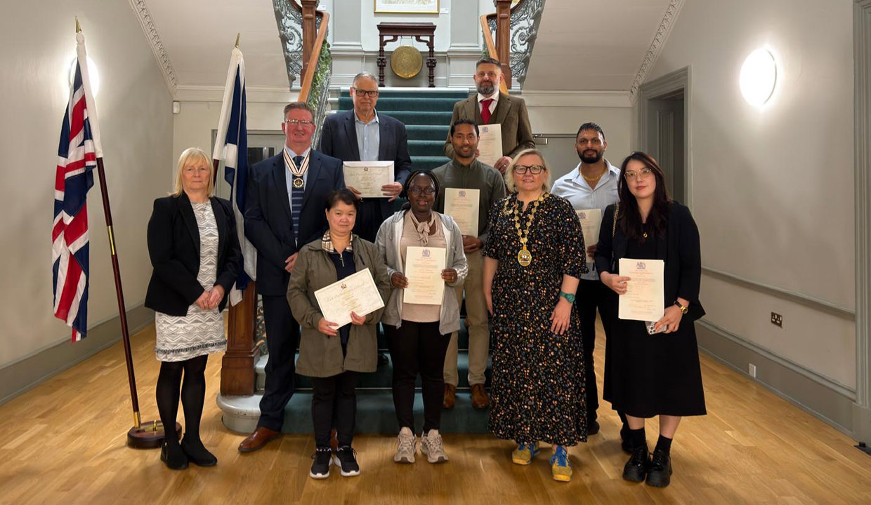 Group of people standing at the foot of sweeping staircase. Behind them are the Union and Saltire Flags. Most of the people are holding certificates. 