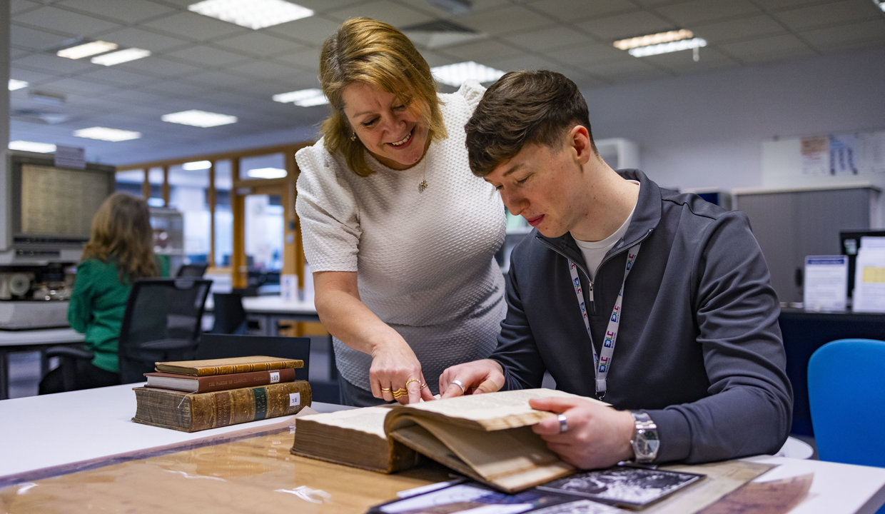 A man sitting at a desk looking at an open book while a woman leans over his shoulder to look at the book. 
