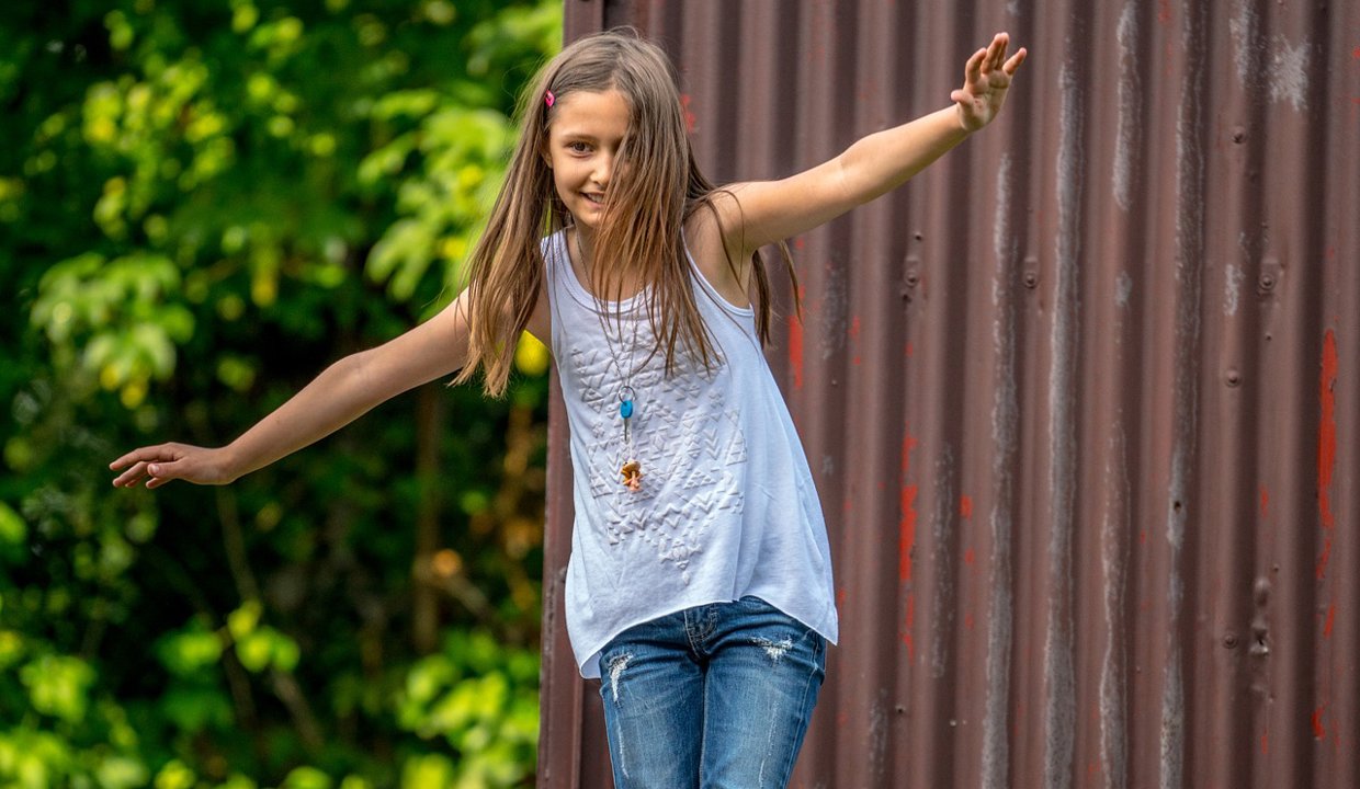 Young girl with long brown hair with her arms out as if to balance herself in front of a corrugated iron wall and green foliage.