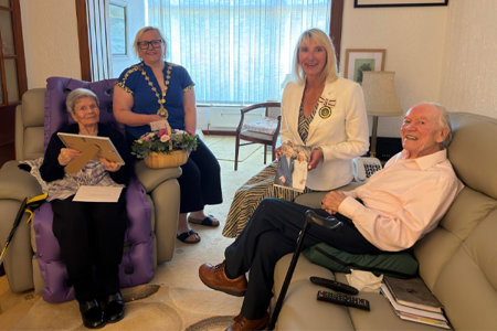 Picture of four people in a living room. On the left is a woman sitting in a chair holding a picture frame.  Next to her is a woman wearing a chain of office and holding a basket of flowers.  Across from them is a woman wearing a medal. She is holding an image of The King and Queen Camilla and she is sitting on the arm of a sofa beside a man.  All four are smiling at the camera. 