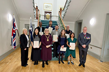 group of people standing at the foot of a beautiful staircase with union and saltire flags on display at the bottom of the stairs on the left hand side. Majority of people in the photograph are holding up their citizenship certificates, Provost Renwick is wearing her chains of office and Deputy Lord Lieutenant Joe Stanfield is also wearing a chain and medals. 