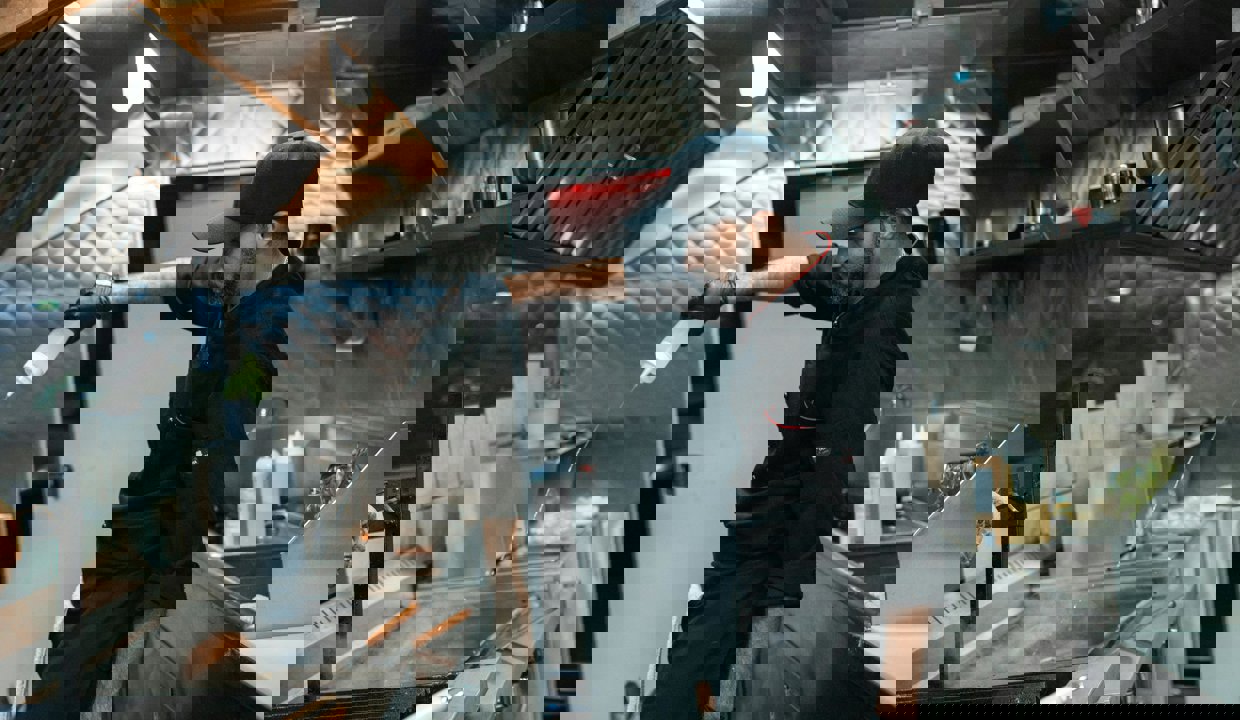 A man dressed in black chef’s clothing and black cap and gloves works at a deep fat fryer in an industrial kitchen